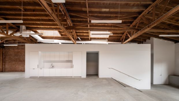 An expansive open-plan interior with a minimalist white kitchen and concealed cabinetry set against exposed red brick walls. The space features a raw concrete floor, white partition walls, and a dramatic exposed timber roof structure with suspended linear fluorescent lighting. A stair or lowered-level opening is visible at right.