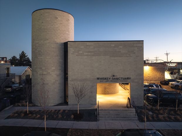 Exterior view of a contemporary masonry building at dusk, featuring a tall cylindrical tower beside a simple rectangular volume. The pale grey brick facade has a large illuminated entrance and signage reading “Whiskey Sanctuary,” with an urban streetscape and parked vehicles surrounding the building.