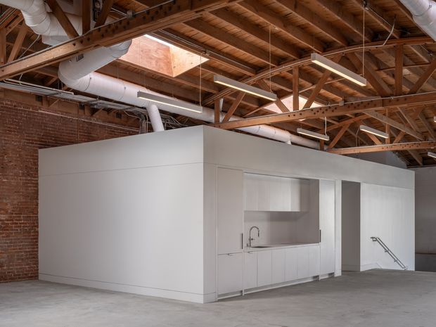 A spacious industrial-style interior with exposed timber roof trusses, brick walls, visible white ductwork, suspended linear lighting, and a polished concrete floor. A minimalist white enclosed service volume contains a compact kitchenette with white cabinetry, open shelving, and a stainless-steel sink. The open-plan space appears suitable for a commercial or creative workspace.