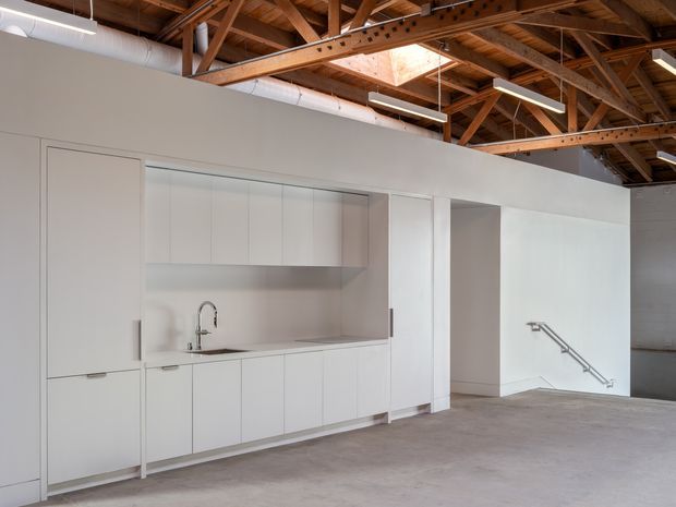 A minimalist commercial or studio interior with a compact white kitchenette built into a white partition wall. The space has polished concrete flooring, exposed timber roof trusses with a skylight, visible ductwork, and industrial-style suspended lighting. A recessed doorway and wall-mounted handrail are also visible.