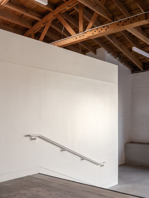 An industrial-style interior with a minimalist white partition wall and accessible handrail, exposed timber roof trusses and decking overhead, white-painted masonry, and a polished grey concrete floor. The space appears to be a converted warehouse or gallery-like circulation area with a stair or ramp concealed behind the wall.