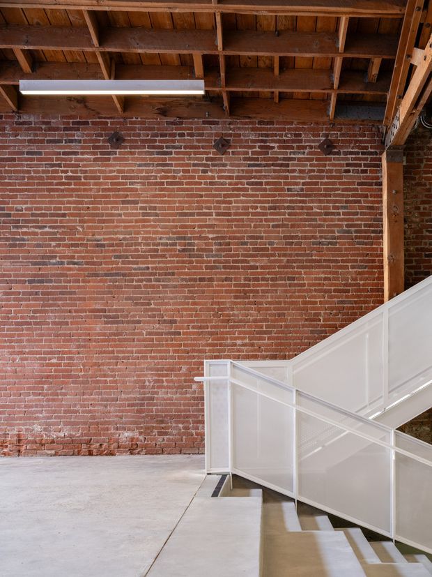 Interior architectural view of an industrial-style space with an exposed timber roof structure, red brick feature wall, polished concrete floor, and a white-painted metal staircase with translucent or solid panel balustrades. A linear fluorescent light is suspended beneath the rafters, creating a strong warehouse or adaptive-reuse character.