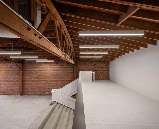 A spacious adaptive-reuse industrial interior with exposed timber roof trusses and beams, original red brick walls, polished concrete flooring, and a bright white mezzanine or upper-level platform. Linear pendant lighting illuminates the open commercial-style space, with stairs and white metal railings connecting the levels.