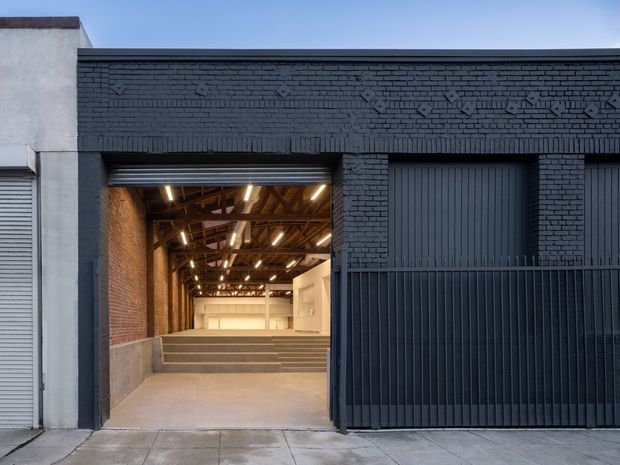 Street-facing view of a converted industrial-style building with a dark painted brick facade, large ribbed metal doors, and an open garage-sized entrance revealing a high-ceilinged interior. The interior features exposed timber roof framing, exposed brick walls, concrete flooring, stepped levels, and linear industrial lighting.