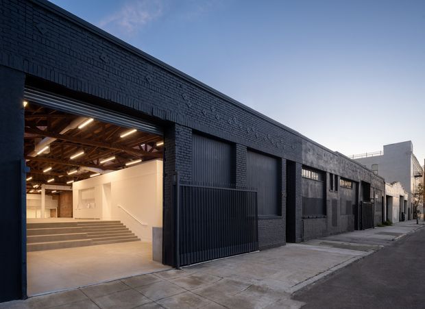 Exterior view of a long, dark-painted brick industrial building on an urban street. The facade features large roller doors, metal gates, recessed openings, and an illuminated open entrance revealing a high-ceilinged interior with exposed timber roof structure and gallery-like steps.