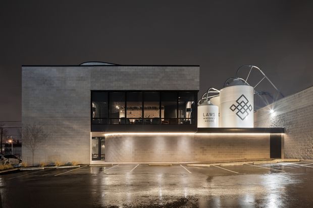 Night-time exterior view of a contemporary commercial building with a rectangular low-rise form, dark-framed upper-level glazing, pale masonry walls, and a strongly illuminated projecting canopy. Large rooftop cylindrical tanks and pipework are prominent, with wet asphalt parking and loading areas in the foreground.
