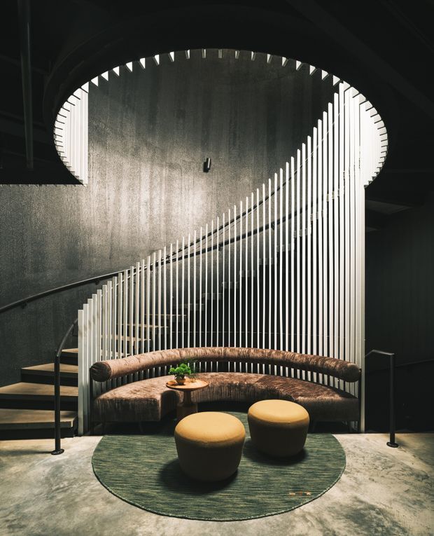 A dramatic, moody interior lobby or lounge featuring a sculptural curved staircase with black treads and slim black handrails. A sweeping white vertical-slat screen follows the stair curve and forms a striking architectural backdrop around a built-in curved upholstered banquette. Two rounded mustard-yellow ottomans sit on a circular dark green rug over a polished grey concrete floor. The space has dark walls, a high curved ceiling form, and strong contrast between black surfaces, white slats, and warm seating.