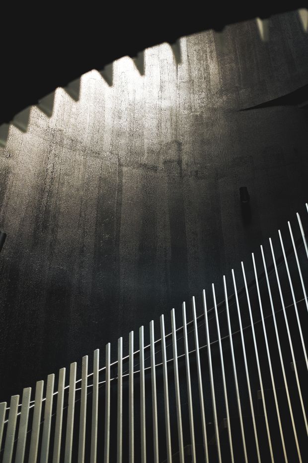 A dramatic upward-looking architectural detail showing a dark, textured concrete or plaster wall with strong light entering through a serrated or sawtooth roof edge. A rhythmic field of slender vertical metal rods forms a railing or screen across the lower portion of the image. The composition is abstract, monochromatic, and highly contrasty, emphasizing shadow, texture, and geometric repetition.
