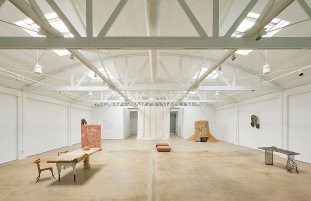 A large, bright white art gallery or exhibition hall with an open-plan layout, exposed painted structural trusses, high ceilings, skylights, and visible ductwork. The polished concrete floor and white walls provide a neutral backdrop for sparse sculptural artworks, plinths, and display tables.