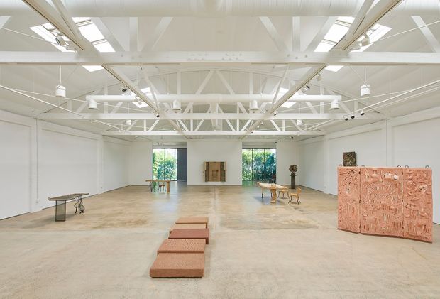 A large, bright white contemporary gallery or exhibition hall with a broad open-plan concrete floor, white-painted exposed steel roof trusses and services, high ceilings, skylights, and tall windows. Sparse artworks and display plinths are arranged across the space, including a large terracotta-toned sculptural panel and low rectangular blocks. The setting has an airy, minimalist industrial character.