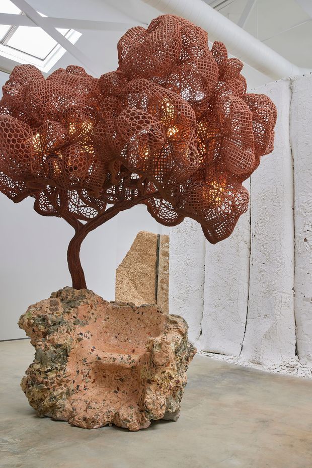 An art-gallery-like interior featuring a dramatic sculptural installation: a rust-red, organic tree form with a dense woven canopy and warm internal lights, rooted in a large rough coral-pink stone or mineral-like sculptural base. The installation stands against white textured walls and a pale polished concrete floor beneath exposed white structural beams, ductwork, and skylights. The image has a highly sculptural, experimental, industrial-art atmosphere.