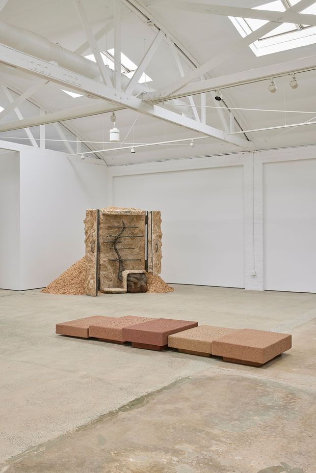 A bright, spacious white-painted industrial gallery or warehouse interior with exposed roof trusses, beams, ducts, track lighting and skylights. The polished concrete floor holds sculptural installations, including a low modular terracotta-toned form in the foreground and a tall textured timber or natural-fibre artwork against the rear wall.