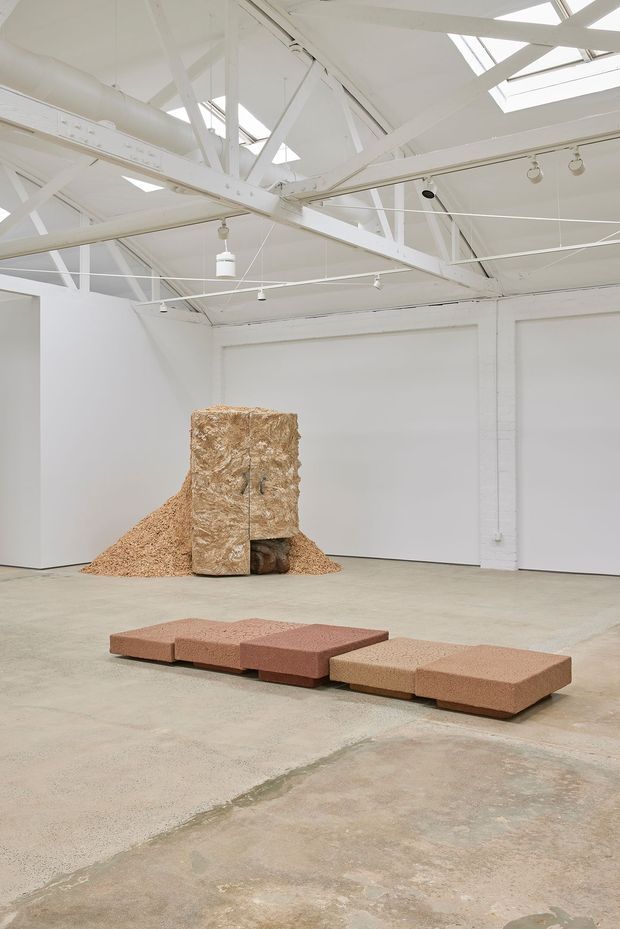 A spacious white-painted industrial gallery interior with exposed structural steel trusses, roof skylights, track lighting, white brick walls, and a worn concrete floor. The room contains a large sculptural installation made from fibrous natural material and a low modular terracotta-toned seating or sculptural arrangement in the foreground.