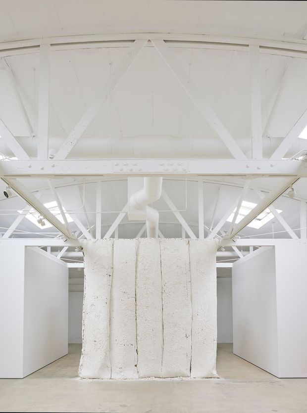 Bright white industrial interior with exposed roof trusses, structural bracing, ductwork, skylights, and a rough central plastered or concrete partition. Large white partition walls create gallery-like openings on either side, with a pale concrete floor and minimal fit-out.