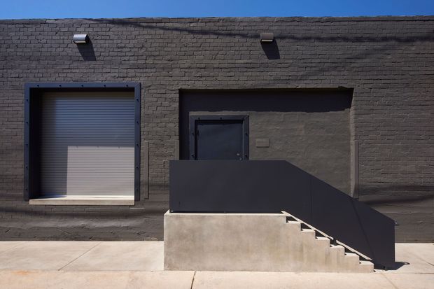 Exterior view of a stark industrial-style brick building facade with a dark recessed entrance, roller garage door, concrete access stairs, and a solid dark metal or painted-steel guardrail. Strong sunlight creates deep shadows across the utilitarian street-facing elevation.