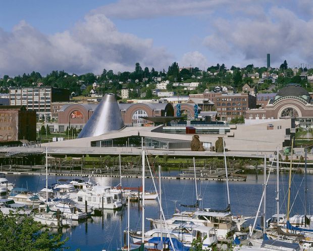 Wide exterior view across a marina toward a prominent waterfront cultural or commercial building with a distinctive curved metallic cone-like roof and broad low-profile forms. The foreground contains numerous moored sailboats and yachts, while a dense hillside neighbourhood and urban buildings rise in the background beneath a partly cloudy sky.