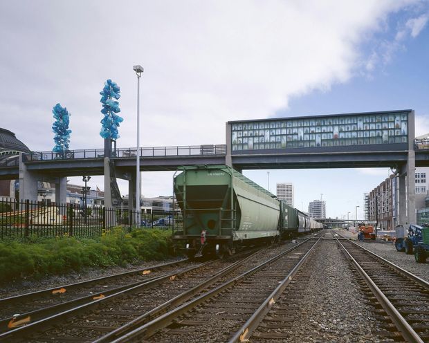 Urban railway corridor viewed at track level, with multiple railway tracks and a green freight train in the foreground. A glazed enclosed pedestrian bridge spans the tracks, with a second elevated bridge behind it, industrial and commercial buildings lining the corridor, fencing, platform-style lighting, and distant city towers. Overcast daylight and utilitarian infrastructure dominate the scene.