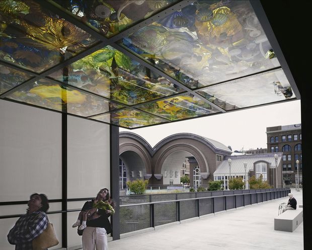 A covered outdoor terrace or elevated walkway beside a historic urban building. The main architectural feature is a dark metal-framed canopy with translucent or glazed panels decorated by colourful botanical and abstract imagery, creating reflections and filtered light overhead. A pale paved floor, dark balustrade, planters, and simple white seating define the space. Beyond the terrace is a prominent masonry building with large arched openings and a nearby ornate light-coloured roof, set among urban buildings.