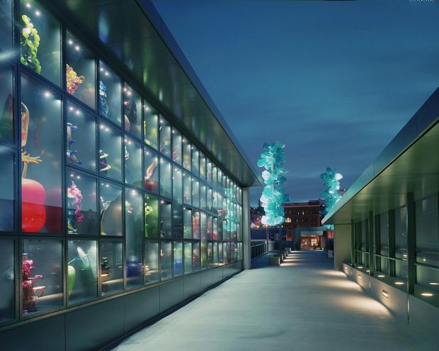 A dusk view of a contemporary commercial complex with a wide illuminated pedestrian passage between two long low-rise buildings. The left building has a dramatic multi-storey glazed display facade with brightly lit sculptural objects inside, while the right building features dark metal or glass panels and recessed linear lighting. Decorative illuminated forms and a building entrance are visible at the far end, creating a polished urban gallery or retail precinct atmosphere.