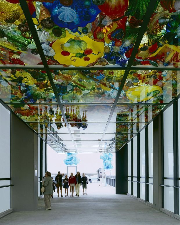 A wide enclosed architectural passageway or gallery with dark structural framing, glass side walls, and a highly distinctive translucent ceiling installation composed of colourful sculptural glass-like forms. Visitors walk through the bright corridor toward an open end, while the ceiling artwork is reflected across the glazing and polished surfaces. The space has a contemporary institutional or commercial character with strong contrast between the neutral structure and vivid overhead artwork.
