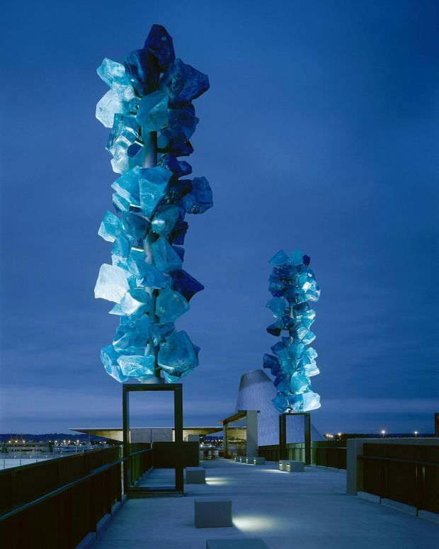 A dusk architectural rooftop terrace or public promenade featuring two towering abstract sculptural installations made from translucent blue, faceted pieces mounted on dark metal frames. The illuminated walkway includes low parapets, benches or plinths, and modern rooftop structures, with a deep blue sky and distant city or waterfront lights beyond.