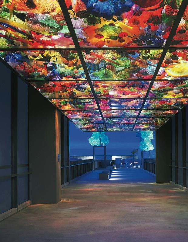 A dramatic interior passage or gallery with dark structural walls and a long illuminated corridor. The ceiling is composed of a black grid supporting an expansive, vividly colourful translucent or printed art installation featuring abstract organic forms. Blue architectural lighting and suspended sculptural elements draw the eye toward a distant open area, while coloured light reflections spread across the polished floor. The space has a highly immersive, exhibition-like atmosphere.