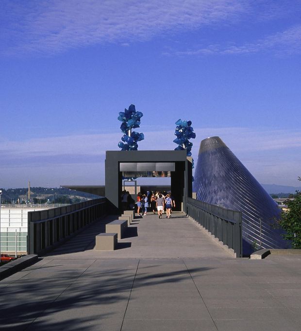 Exterior architectural photograph of a broad elevated pedestrian approach leading to a dark-framed entrance structure, with a distinctive curved, ribbed metallic roof or building form rising to the right. People are visible near the entrance, with railings, paved surfaces, and a distant urban landscape beneath a blue sky.
