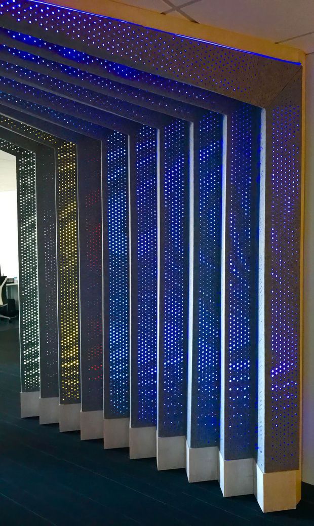 Interior architectural feature showing a series of tall, perforated dark panels or fins arranged in a repeating corridor-like rhythm, illuminated with blue LED points and strips. The panels appear to form a decorative partition or screen, with a dark floor and a small glimpse of an office-like area beyond the opening. The ceiling is a simple suspended grid.