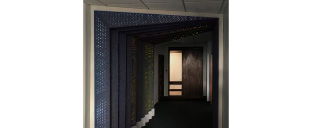 A dark, atmospheric interior corridor framed by multiple deep door-like portals or wall panels. The layered frames are decorated with dense small perforations or pinpoints of light, creating a dramatic tunnel effect toward a softly illuminated doorway at the far end. The ceiling has a plain suspended grid, while the corridor floor and walls are predominantly dark.
