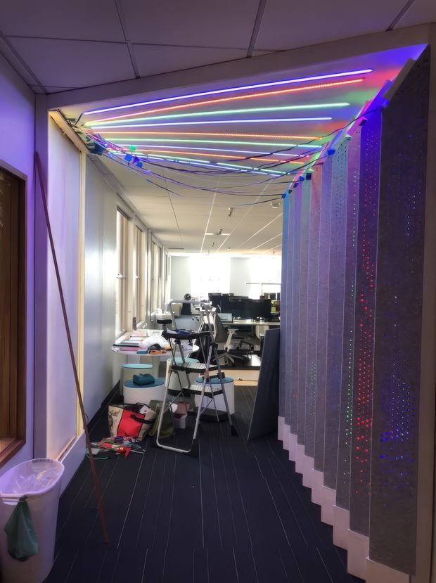 A narrow contemporary commercial interior or office corridor leading into an open-plan workspace. The space features dark flooring, white walls and a suspended grid ceiling, with desks, computer monitors and office chairs visible at the far end. The corridor is dominated by a colorful installation of exposed multicolored linear LED lights and hanging wiring across the ceiling, alongside tall dark perforated or translucent partition panels with colored pinpoints of light. The overall appearance is functional but highly theatrical and technology-oriented.