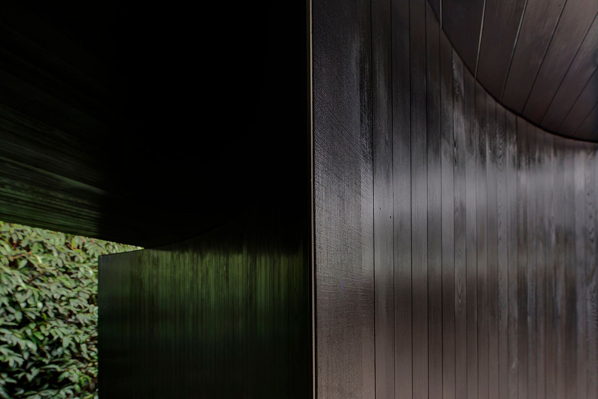 The dark toned cladding absorbs the colour tones of the adjacent spaces; the green of the hedge and the grey/brown of the gravel courtyard.