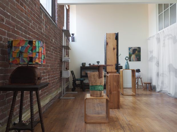 A bright, high-ceilinged loft-style interior with exposed red-brick walls, wide timber floorboards, and large industrial-style windows. The room functions as a combined living, dining, and workspace, with sculptural timber furniture, a desk and chair at the rear, a small dining table and stools, artwork, and a colourful tiled or fabric-covered wood-fired oven structure along the left wall. Sheer curtains soften the tall glazing on the right.