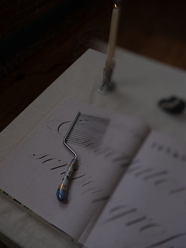 A dimly lit close-up of an open book or sketchbook on a light tabletop, with handwritten marks, a small metal rake-like tool resting across the pages, and a tall lit candle in a holder. The scene has a moody, intimate still-life quality, with the surrounding room mostly obscured in shadow.