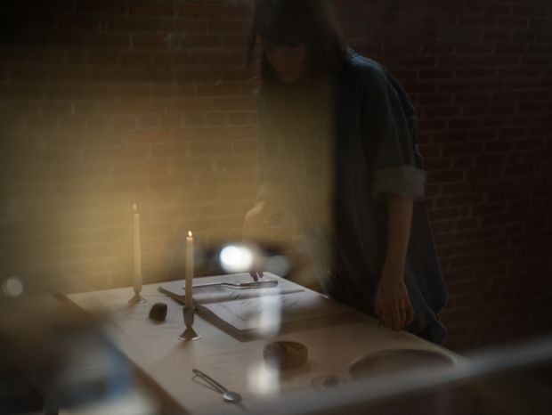 Moody, softly lit interior scene with a person standing at a dining table set with candles, plates, cutlery and bread. Exposed dark red brick walls form the backdrop, while warm candlelight and shallow-focus reflections create an intimate atmosphere.