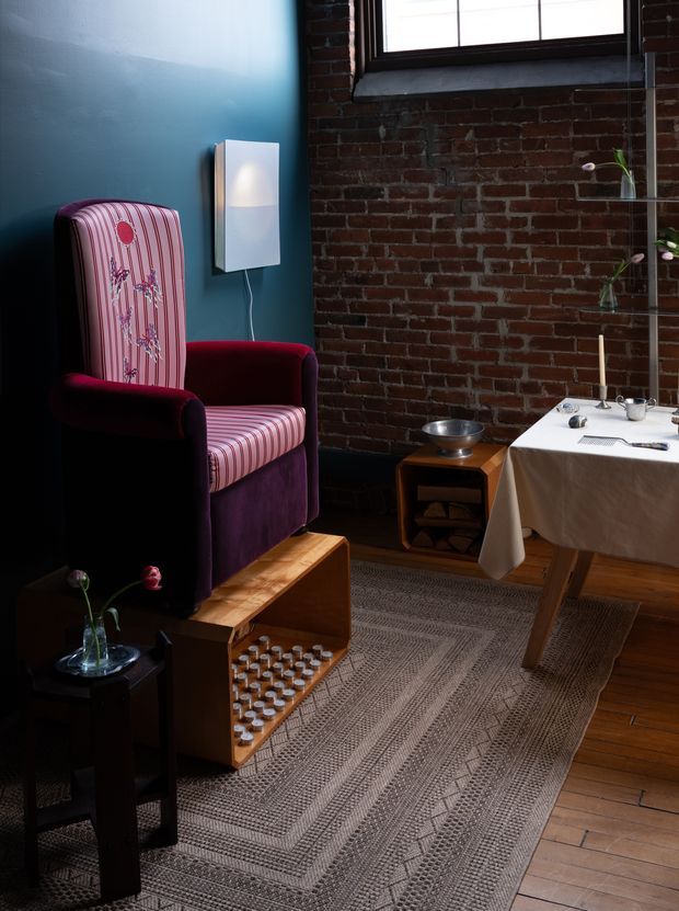 A moody, eclectic living area with an upholstered wingback chair in deep purple and pink stripes, a small wooden footstool, patterned neutral rug, dark timber flooring, exposed red brick walls, and a blue painted accent wall. A simple wall light, side tables, and a dining or work table with a white cloth complete the space. Natural light enters through a dark-framed window.