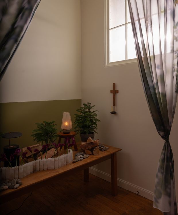 A softly lit interior corner featuring a timber console table styled with potted plants, pebbles, a glowing table lamp, and a small wooden cross mounted on the wall. A tall divided-pane window with sheer curtains brings in muted daylight. The lower wall is painted olive green while the upper wall is warm off-white, and the floor is dark timber.