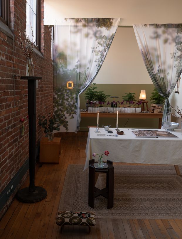 A warm, intimate dining room in a converted-looking brick interior, with exposed red brick walls, timber flooring, gauzy botanical curtains, a long dining table set with candles and tableware, indoor plants, and layered woven rugs. A black freestanding fireplace and soft ambient lamps add a cozy, eclectic atmosphere.