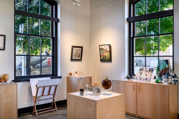 Bright, contemporary gallery-like interior with tall black-framed multi-pane windows, white walls, dark trim, and light timber display cabinetry. A central light-wood island or plinth displays decorative objects, while framed artwork, prints, and small furnishings reinforce the showroom or studio character. Leafy trees and neighboring buildings are visible through the windows.