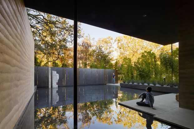 A dark, minimalist architectural courtyard with a shallow reflective pool extending between textured concrete walls and a covered terrace. Floor-to-ceiling glazing or open structural framing creates strong reflections of surrounding autumn trees, while a child sits on a projecting paved platform beside low planted beds.