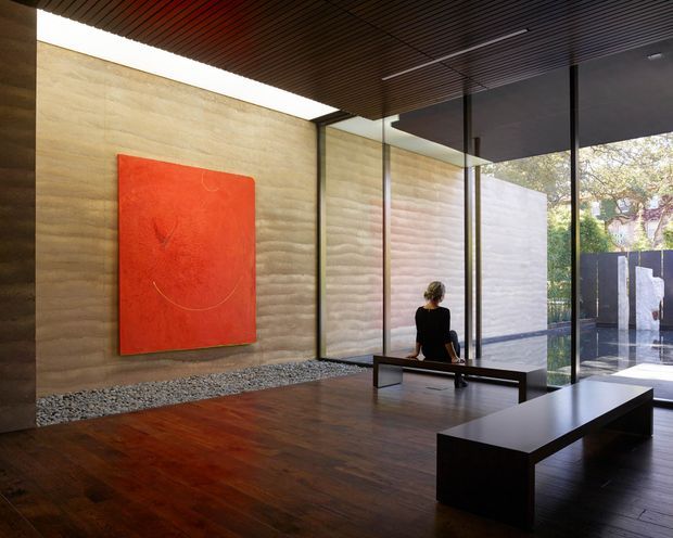 A refined modern interior, likely an entry foyer or gallery-like living space, with a richly stained timber floor, sculptural black bench seating, a long dark slatted ceiling, and full-height glazing. Textured horizontal concrete walls frame a large red abstract artwork, while a landscaped exterior courtyard is visible through the glass.