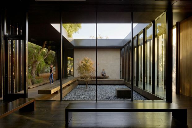 A dark, refined modern interior opens onto a minimalist enclosed courtyard through extensive floor-to-ceiling glazing. The space features dark timber floors and ceilings, black-framed glass walls, built-in timber seating and low joinery, a gravel courtyard with a shallow reflective pool, and a strong indoor-outdoor connection.