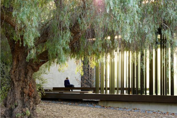 A tranquil landscaped courtyard or outdoor garden space beneath a mature, spreading tree. A long dark timber bench and low platform sit beside a contemporary vertical slatted screen, with filtered sunlight and dense greenery creating a secluded, contemplative setting.