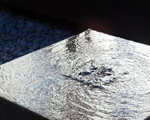 Close-up abstract view of a square water feature or basin surface, with rippling reflective water catching bright light. Dark surrounding edges and a bed of blue-grey stones are visible in the background.