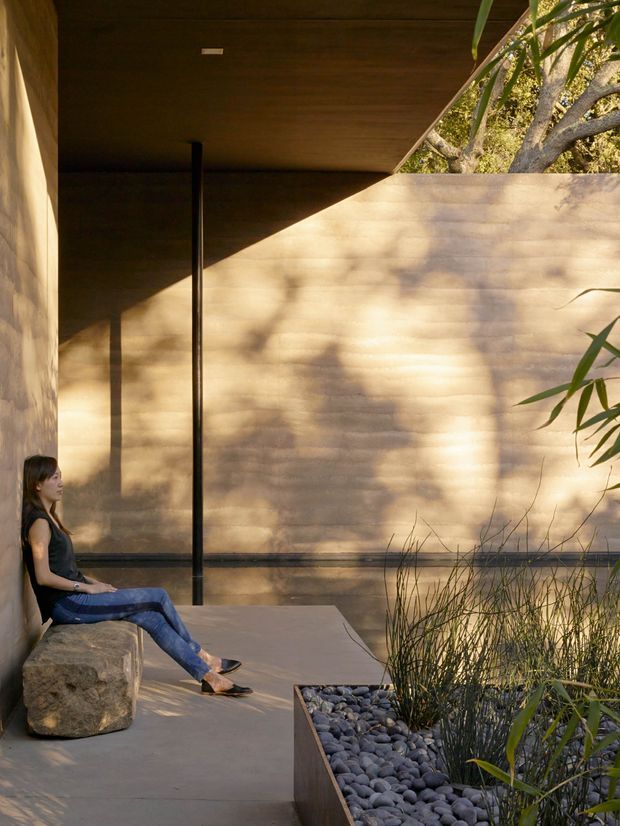 A serene, minimalist covered outdoor courtyard or terrace with a warm wood-lined ceiling, smooth tan rendered walls, dark slender structural posts, and a concrete floor. A raised planter filled with dark river stones and ornamental grasses borders the space, while a woman sits on a large natural stone bench at the left. Strong sunlight and tree shadows create a calm connection between the sheltered architecture and surrounding landscape.