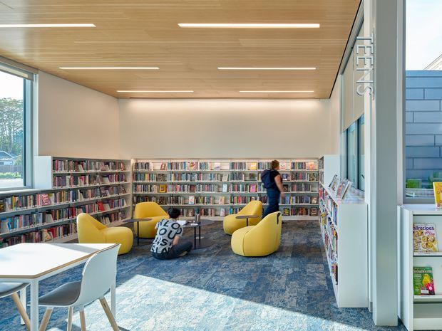 A bright contemporary library reading area with floor-to-ceiling glazing, extensive wall-mounted bookshelves, colorful upholstered lounge chairs, small reading tables, and a patterned blue-grey carpet. The space features a warm timber slatted ceiling with recessed linear lighting and a clean white interior fit-out. Magazine shelving and the vertical “TEENS” signage are visible along the glazed side.