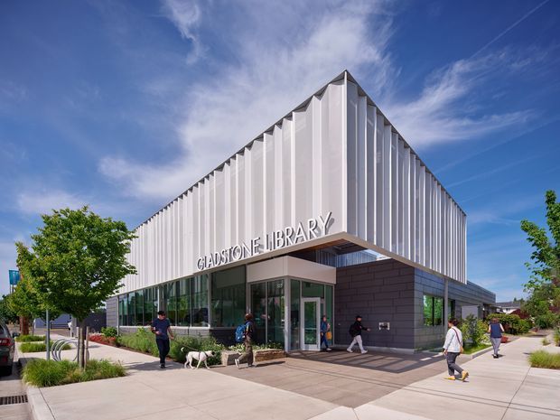 Exterior view of the Gladstone Library, a contemporary civic building with a sharply pitched, folded roof form and prominent vertical white facade fins. The glazed entrance is set beneath a projecting canopy, with dark masonry walls at the lower level and landscaped pedestrian paving around the building.