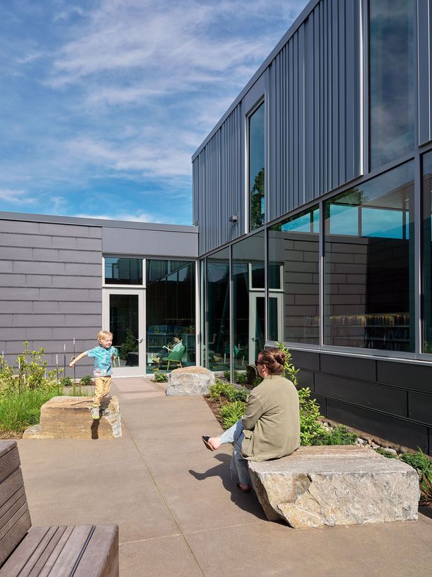 Exterior view of a contemporary low-rise building arranged around a landscaped courtyard. The facade combines dark grey horizontal masonry or panel cladding with vertically ribbed dark metal cladding, extensive black-framed glazing, and a low flat roof. A paved pedestrian courtyard includes large natural stone seating blocks, planting beds, and timber bench seating. People are present, and interior library-like shelving and seating are visible through the windows.