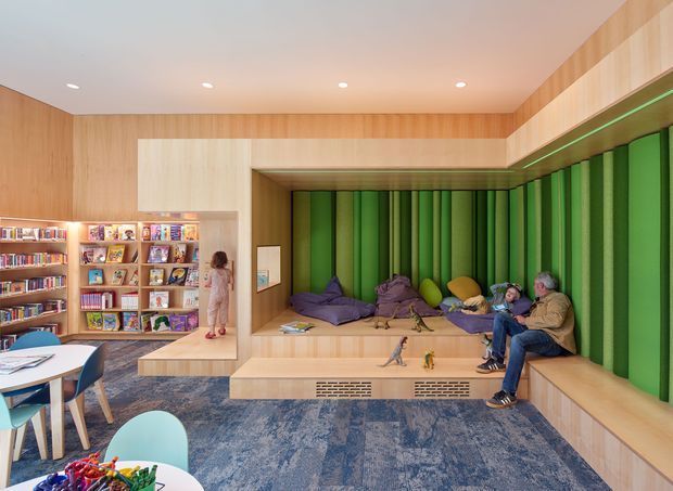 A brightly designed library or reading lounge with extensive built-in timber bookshelves, a large recessed window seat, and vivid green vertical padded wall panels. The space includes integrated bench seating with cushions, adults and children using the reading area, a small table and chairs, recessed ceiling lights, and blue patterned carpet. The architecture combines warm timber lining and joinery with a playful, contemporary interior fit-out.