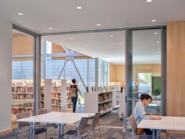 Bright contemporary library reading area with open shelving filled with books, large glazed windows, white study tables, chairs, and a visible student workspace. The space has a clean neutral interior, warm timber wall panelling, exposed structural glazing supports, and abundant natural light.
