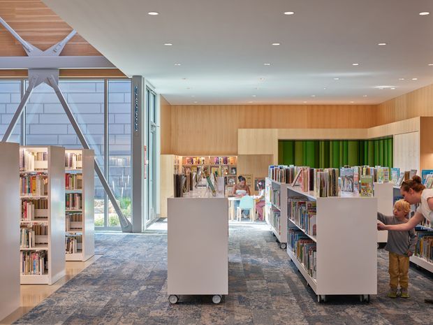 Bright contemporary library interior with rows of bookshelves, mobile white display units, a patterned carpet floor, and extensive glazing. Warm timber wall and ceiling finishes contrast with white shelving and exposed structural diagonal braces at the window wall. Children and an adult are browsing the collection.